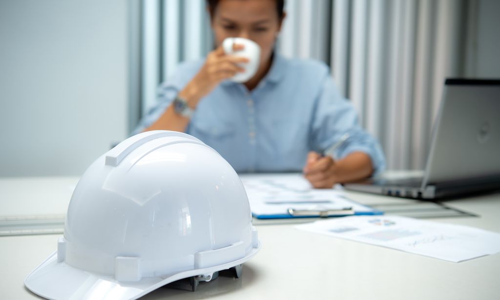 Ein Schutzhelm liegt auf einem Tisch. Im Hintergrund sitzt eine Person an diesem Tisch, schreibt etwas auf ein Papier und trinkt Kaffee. 