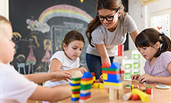  Junge Kindergartenhelferin beaufsichtigt die Kinder in einem Kindergarten beim Spielen mit Bauklötzen. 